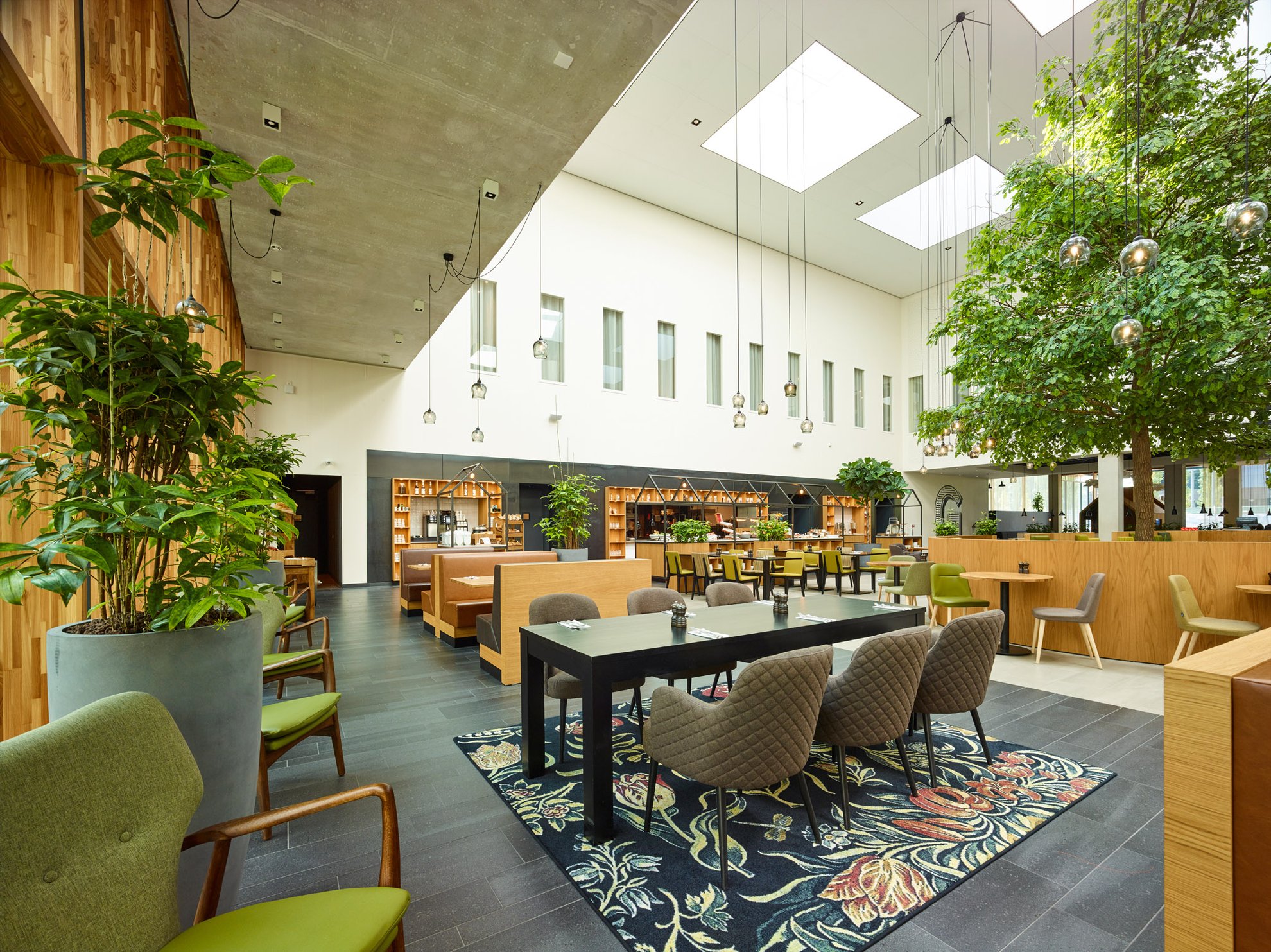 Modern hotel lobby restaurant with natural light, indoor greenery, wooden seating areas, and communal dining tables